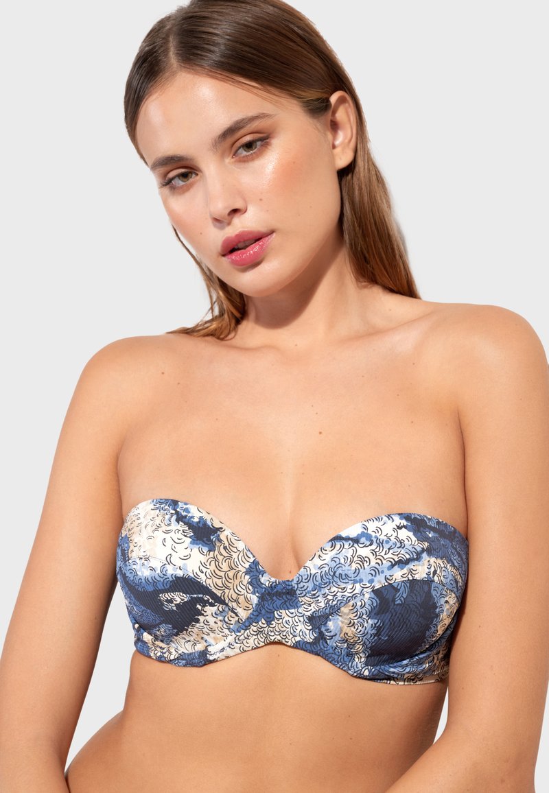 Femme aux cheveux raides châtain clair portant un haut de bikini bustier bleu et beige à motifs, sur un fond gris clair uni.