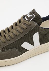 Sneaker verde oliva com tecido texturizado, detalhes em branco no logotipo e sola de borracha bege. Apresenta fecho de atacadores e língua almofadada.