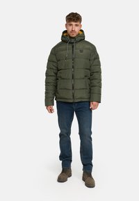 Groene pufferjas met een rits, verstelbare capuchon, grijze trekkoorden en gele voering, gecombineerd met blauwe jeans en bruine laarzen.