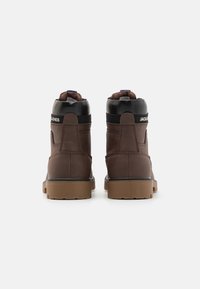 Botas de cuero marrón hasta el tobillo con acentos en negro, que presentan una suela texturizada, paneles laterales elásticos y una etiqueta de marca en la parte posterior.