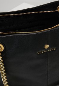 Sorte læderhåndtaske med en guld kæderekke, lynlåslukning og præget "GUESS LUXE" logo. Indvendigt har den stofforet med logo-mønster.