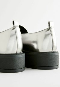 Zapatos metálicos plateados con una superficie superior lisa y una suela negra texturizada. Presentan un tacón bajo y un diseño recortado en la parte trasera.
