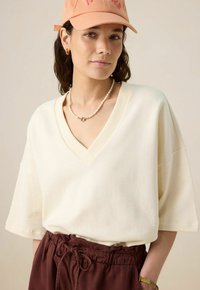 T-shirt oversize  à col en V crème avec des manches courtes, associé à un pantalon bordeaux à cordon de serrage. Les accessoires comprennent un collier en perles et une casquette pêche.