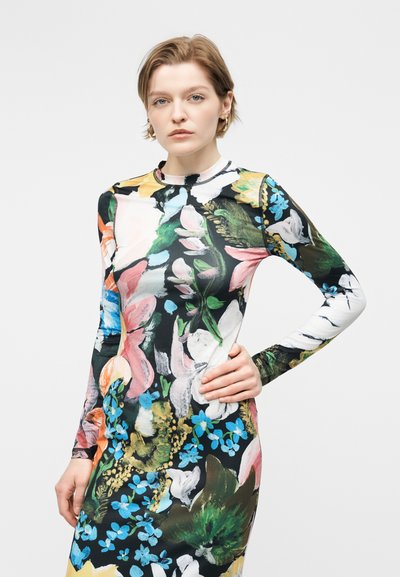 Γυναίκα με κοντά μαλλιά που φοράει ένα μακρυμάνικο, ψηλό λαιμό φόρεμα με πολύχρωμο λουλουδάτο σχέδιο σε μαύρο φόντο.