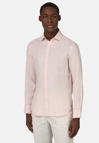 Camicia in lino rosa chiaro con colletto a button-down e maniche lunghe. Presenta una leggera texture e orlo arrotondato, abbinata a pantaloni chiari.