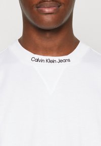 Sudadera de cuello redondo blanca con el texto negro "Calvin Klein Jeans" en el cuello, hecha de material de algodón suave con una textura lisa.