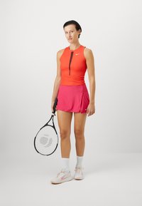 Piros ujjatlan sporttop ribbesített textúrával és fekete cipzárral, rózsaszín szoknyával párosítva. Fekete teniszütőt tart, fehér zoknikat és cipőket visel.