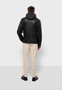 Chaqueta de cuero negra con capucha, acabado texturizado y un diseño ajustado, combinada con pantalones beige claros y zapatos negros.