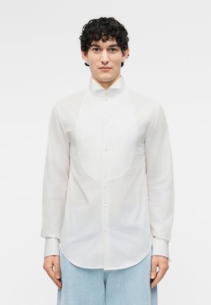 Młody mężczyzna z kręconymi włosami, ubrany w białą koszulę zapinaną z przodu na guziki i jasnoniebieskie jeansy, stojący na jednolitym tle.