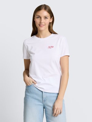 Jeune femme aux longs cheveux bruns portant un t-shirt blanc avec un texte rouge et un jean bleu clair, debout avec une main dans la poche.