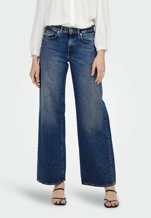 Kvinde iført højtaljede, vidde blå jeans kombineret med en hvid bluse med lange ærmer og sorte åbne hæle, stående mod en ensfarvet baggrund.