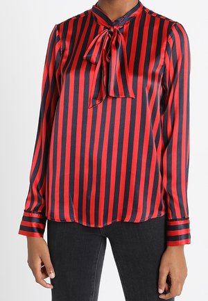 Femme portant un chemisier rouge et noir à rayures verticales à manches longues, avec un nœud devant, associé à un pantalon noir.