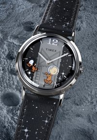 Runde Timex-Uhr mit einem schwarzen Lederarmband, das ein sternenförmiges Muster aufweist; Grafik von Astronauten auf einem dunklen Zifferblatt mit silbernen Stundenmarkierungen.