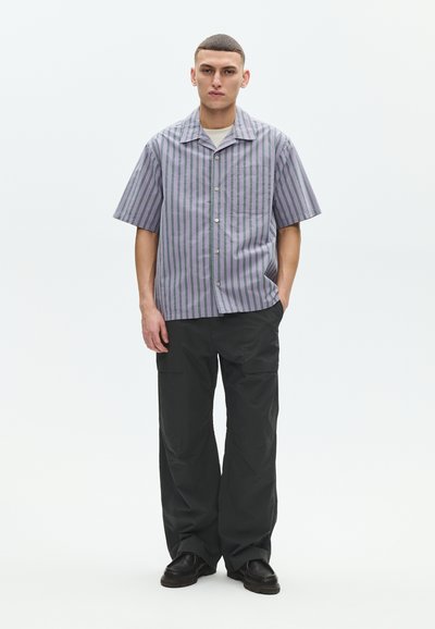 Νέος άντρας που φοράει κοντομάνικο πουκάμισο με κουμπιά και ρίγες, μαύρο παντελόνι με φαρδιά πόδια και μαύρα παπούτσια, στέκεται με το ένα χέρι στην τσέπη.