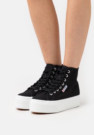 Baskets hautes en toile noire avec semelles épaisses blanches à plateforme, portées sur des jambes nues, avec des lacets sur le devant et des étiquettes avec le logo de la marque.