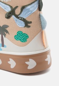 Κοντινό πλάνο πτέρνας παπουτσιού με κέντημα δεινοσαύρου, πράσινο γραφικό δέντρου και καφέ σόλα με λευκά αποτυπώματα δεινοσαύρου.