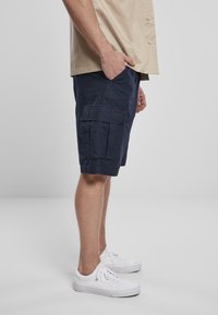 Shorts cargo navy avec plusieurs poches, fabriqués en tissu léger. Associés à des baskets blanches. Le cadre est minimaliste et neutre.