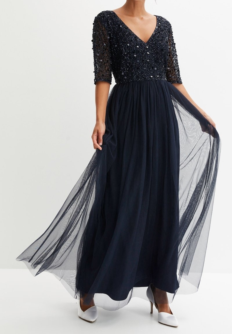 Femme portant une longue robe bleu marine avec un haut orné de perles et une jupe fluide transparente, associée à des chaussures blanches à talons hauts, tenant les côtés de la jupe.