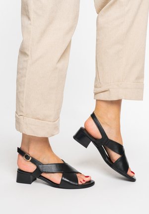 Feet wearing black low-heeled leather sandals with ankle straps and beige cuffed pants against a white background.