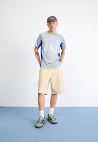 Light grey athletic shirt with blue accents and white text, paired with beige plaid shorts. Black and green sneakers, white socks.
