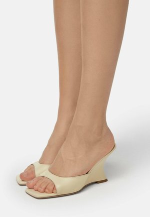 Beige leather wedge sandals with an open toe design, featuring a wide band across the midfoot and a sculpted heel. Smooth texture.