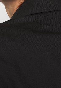 Nära bild av svart tyg med en texturerad yta, med en synlig sömdetalj vid kragområdet.