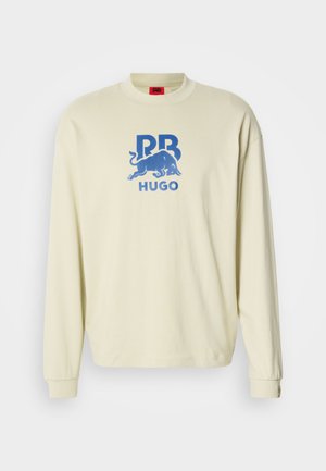 Svetr krémovej farby s dlhými rukávmi, okrúhlym výstrihom a modrou grafikou zobrazenou na prednej strane, ktorá obsahuje býka a text "HUGO". Mäkká textúra.