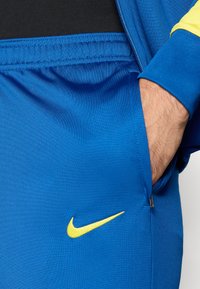 Μπλε αθλητικό μπουφάν από ελαφρύ ύφασμα, με κίτρινο λογότυπο Nike στο στήθος και εσωτερικό σε αντιθετικό μαύρο χρώμα. Περιλαμβάνει τσέπη με φερμουάρ.