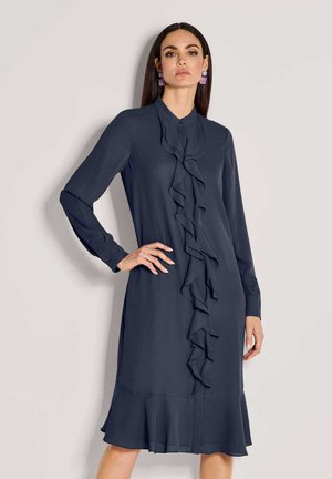 Frau posiert in einem dunkelblauen, langärmeligen Kleid mit Rüschen vorne und am Saum, trägt violette Tropfenohrringe vor hellem Hintergrund.