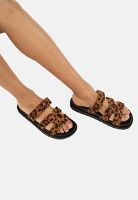 Sandali marroni con stampa leopardata, dotati di due fasce regolabili, suola nera piatta e design della punta arrotondata. La texture appare morbida e simile alla pelle scamosciata.