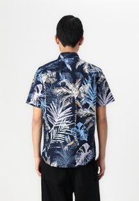 Chemise à manches courtes bleu marine avec un imprimé de feuilles tropicales et d'oiseaux dans des tons de bleu clair et de crème, présentant une coupe décontractée et un ourlet courbé.