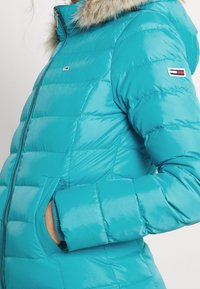 Turquoise gewatteerd jack met capuchon gevoerd met bont, ritssluiting en elastische manchetten. Heeft een logoaccent op de linkermouw en een doorgestikte textuur.