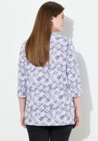 Lyse lilla top med blomsterprint i sort og hvid, med trekvartærmer og en løs pasform. Sorte bukser er synlige nedenfor.