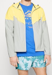 Muž oblečený v barevně rozdělené žluté a šedé bundě, modrém tričku s potiskem a modro-zelených šortkách se vzorem, stojí se uvolněnýma rukama.