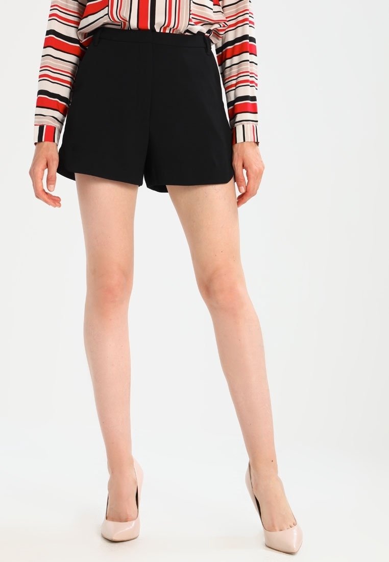 Shorts noirs sur mesure avec une texture lisse, présentant une coupe droite et des lignes nettes. Associés à un chemisier rayé multicolore et des talons clairs.