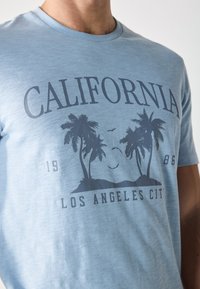 Svijetloplavi pamučni majica s grafičkim dizajnom palmi i natpisom "CALIFORNIA" u tamnoplavoj boji, okrugli izrez, kratki rukavi.