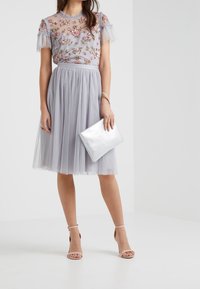 Vestido de tul gris claro con bordado floral, mangas cortas con volantes y cintura ajustada. Combinado con un bolso de mano plateado y tacones nude.