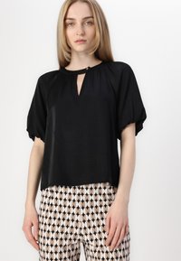 Black short-sleeve top with a keyhole neckline, flowing silhouette, and soft texture, worn with patterned pants featuring an oval design.