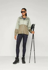 Ľahká dvojfarebná bunda v olivovej a béžovej farbe, čierne legíny, robustné čierne čižmy a slnečné okuliare. Drží turistické palice.