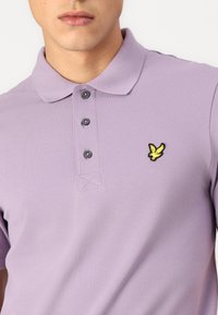 Lilla polo-skjorte laget av teksturert stoff med krage, tre knapper og et gult og svart fugleemblem på venstre bryst.