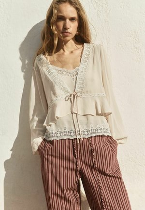 Tânără purtând o bluză bej cu dantelă la margină, cu mâneci lungi, și pantaloni largi maro cu dungi, stând în picioare lângă un perete alb.