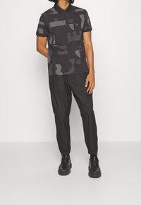 Černé polo tričko s šedými geometrickými vzory, krátké rukávy a límeček. V kombinaci s černými zúženými kalhotami a černými šněrovacími botami.