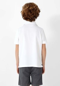 Camiseta polo blanca de manga corta con cuello, tela texturizada, vista trasera, combinada con shorts grises, con aberturas laterales en el dobladillo para mayor comodidad.