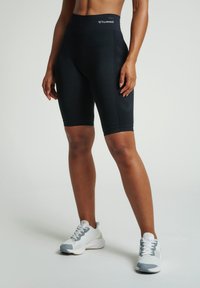 Osoba v černých sportovních šortkách s vysokým pasem značky Hummel a bílých-šedých teniskách stojí na bílé podlaze.