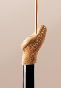 Applicatore di fondotinta con una morbida punta a pennello beige, attaccato a un elegante tubo nero. Gocce di fondotinta liquido scorrono dalla punta.