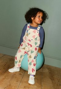 Svetlo kremna kombinezona s pisanimi vzorci src, združena z modro dolgoročno majico in belimi čevlji, sedi na modri žogi.