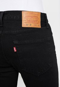 Jeans de mezclilla negra con un parche trasero de cuero que presenta texto y gráficos en relieve. Etiqueta roja de Levi's en el bolsillo lateral, diseño clásico de cinco bolsillos.