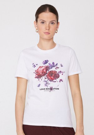 Kobieta w białej koszulce z czerwonymi i fioletowymi kwiatami oraz tekstem "LOVE REVOLUTION WHISPERS OF GRACE", zwrócona twarzą do przodu na białym tle.