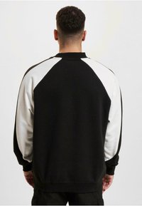 Sweat-shirt noir et blanc avec un design raglan. Col et poignets côtelés, texture douce. Corps noir, manches blanches avec des détails de coutures contrastants.