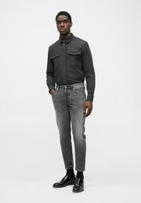 Camicia di jeans nera con due tasche sul petto, abbinata a jeans grigi a gamba affusolata e mocassini neri, su uno sfondo neutro.